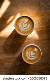 Coffee on wooden background. Two cups of cappuccino with latte art on brown table with sunbeam. Fresh morning coffee with delicious milk foam. Top view, flat lay. - Shutterstock ID 2005058393