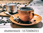 Coffee in an orange cup color. Milk in a metal cup.  Coffee in the side light. Spilled coffee beans. Still life in vintage style or in rustical style. Coffee on a pale oak table.