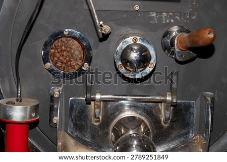 Coffee roaster machine control panel with sight glass window and sample spoon close up. Industrial drum roasting machinery faceplate with brown arabica beans during manufacturing process, technical close-up.