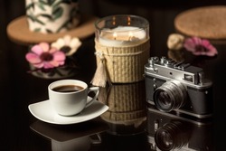 Coffee Scene With Vintage Camera. A Cup Of Coffee And Vintage Photo Camera On A Dark Glass Table With 

Candles And Flowers. Still Life Concept With Coffee And Old Camera