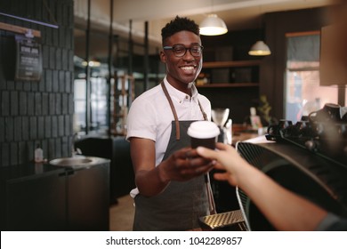 Coffee Shop Owner Handing Over A Sealed Coffee Cup To A Customer. Man Serving Customer With A Smile At A Coffee Shop.