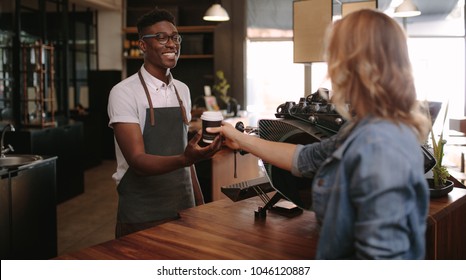 Coffee Shop Owner Handing Over A Sealed Coffee Cup To A Customer. Man Serving Customer With A Smile At A Coffee Shop.