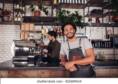 Coffee shop worker smiling to camera, standing at the counter. Happy young man in apron and hat leaning to cafe counter, with waiter working in background. - Powered by Shutterstock