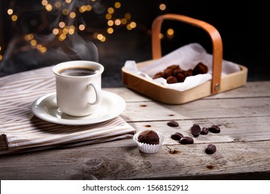 Coffee table setting with chocolate sweets and steaming cup of coffee on rustic wooden table with garland as background