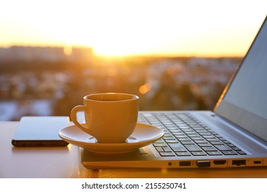 Coffee Or Tea Cup On Laptop Against The Window With Sunshine. Cozy Workplace In Home Office, Inspiring View To Morning City