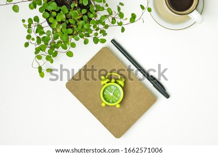 
Coffee time. Top view on a white table with a notebook with dark sheets, pen, green alarm clock, indoor flower, cup of coffee. Time to drink coffee, coffee break, Working day concept, flat lay