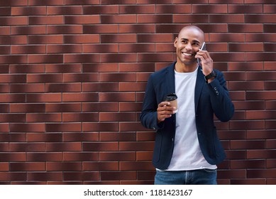 Coffee time. Young guy standing isolated leaning back on wall holding cup drinking coffee talking on smartphone smiling happy