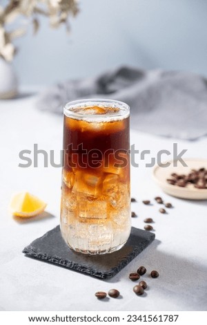 Coffee with tonic in a tall glass with ice and lemon on a light blue background with coffee beans and fruit. Trendy summer craft refreshing drink. Front view.