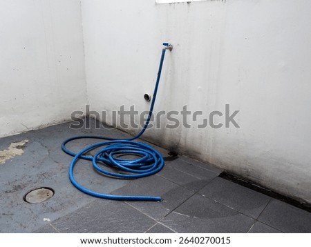 A coiled blue garden hose lies on a grey concrete floor, connected to a wall-mounted water spigot. useful for concepts of water conservation, home maintenance, gardening, or industrial cleaning.