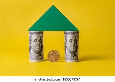 A coin in denomination of 1 American dollar under a green wooden roof of a conventional house, the walls of which consist of 100 USD bills rolled in a tube, on a yellow background