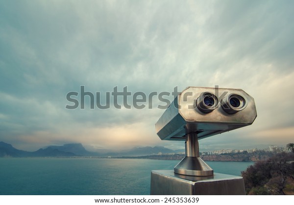 Coin Operated Binocular\
viewer next to the waterside promenade in Antalya looking out to\
the Bay and city. Landscape with beautiful cloudy sky, sea and\
mountains..