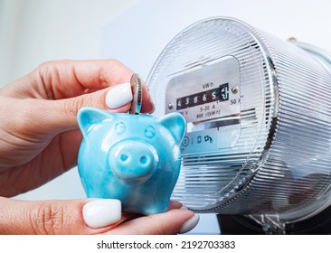 Coin And Piggy Bank In Woman's Hands Near Electricity Meter At Home. Symbolic Image Of Energy Costs And Electricity Prices, Saving The Household Budget On Paying Utility Bills.