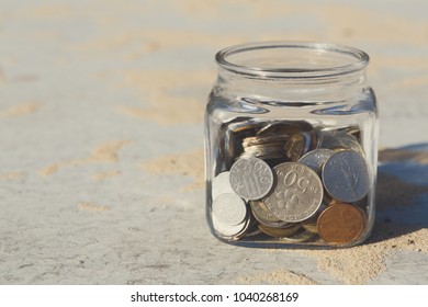 Coins In Glass Jar On Gray Floor With Sand Background, Copy Space. Money Box, Saving Money For Dream, Pension, Vacation. Financial Stability Concept