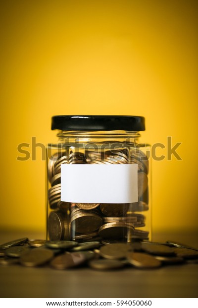 A lot coins in glass money jar with blank\
white paper on yellow\
background.