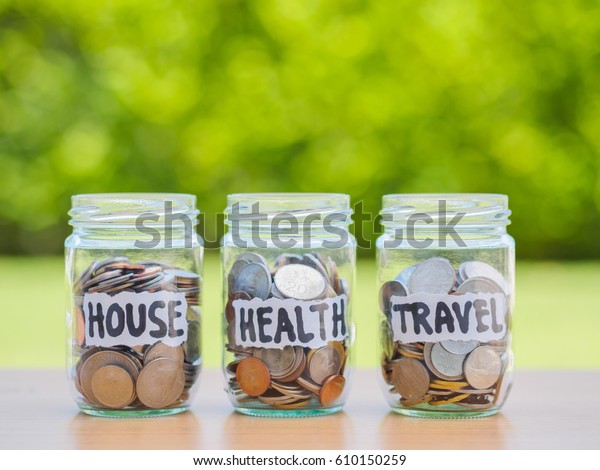 A lot coins in glass money jar\
on the wood table. Saving for house, health and travel\
concept.