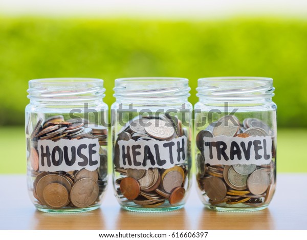A lot coins in glass money jar\
on the wood table. Saving for house, health and travel\
concept.