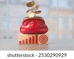 Coins are spilling out of a red purse atop wooden blocks that display financial graphs and a target symbol, suggesting a focus on economic success and savings.