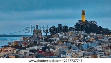 Coit Tower aerial at dusk with lights on in buildings and view of Oakland Bay Bridge