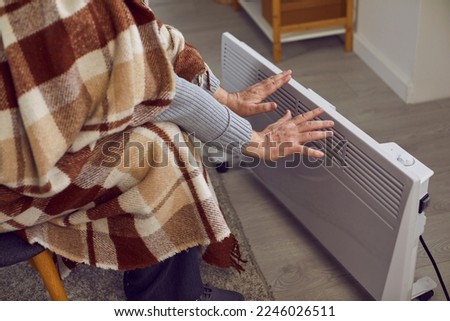 Cold in apartment. Senior man at home warms his hands on electric heater due to problems with central heating. Cropped image of pensioner wrapped in woolen plaid holding his hands close to heater.