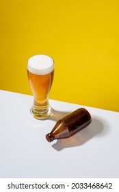 Cold Beer Served In A Glass With White Foam, An Empty Glass Bottle On One Side, On A White Table And Yellow Background, No People