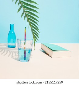 Cold Blue Drink In Glass And Pastel Blue Book Under Green Palm Branch. Palm Tree Shadow Imitating Beach Summer Day. Aesthetic Abstract Design Idea. Sunny Beach Day Concept.