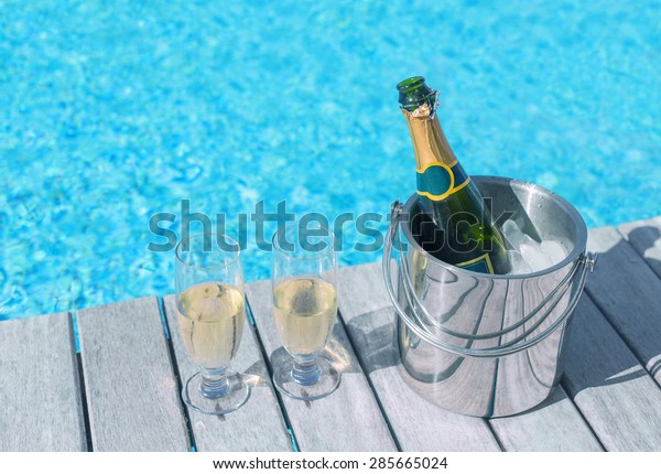 Cold champagne bottle in ice\
bucket and two glasses of champagne on the deck by the swimming\
pool