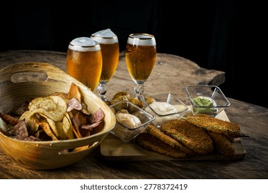 Cold draft beer served with crispy snacks and dipping sauces on rustic wooden table against dark background. Assorted potato chips, fried appetizers, and creamy dips create a cozy pub atmosphere perfect for bar, brewery, and party concepts.