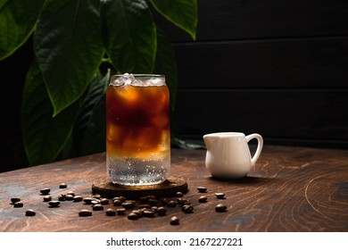 Cold espresso tonic. A highball glass filled with ice cubes, tonic soda water, coffee beans around, milk jug, wooden background