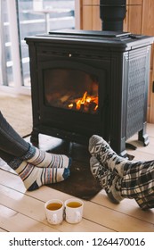 Cold Fall Or Winter Day. People Drinking Tea And Resting By The Stove. Closeup Photo Of Human Feet In Warm Woolen Socks Over Fire Place. Hygge Concept Of Cozy Winter Weekend In Cabin.