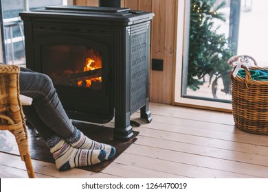 Cold Fall Or Winter Day. Woman Resting By The Stove. Closeup Photo Of Human Feet In Warm Woolen Socks Over Fire Place. Hygge Concept Of Cozy Winter Weekend In Cabin.