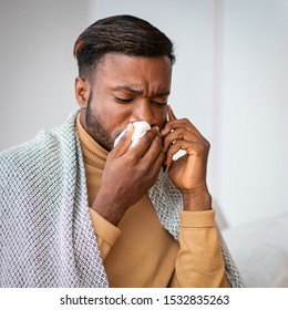 Cold And Flu. African American Man Blowing Runny Nose In Paper Tissue Talking On Phone Wrapped In Blanket At Home.