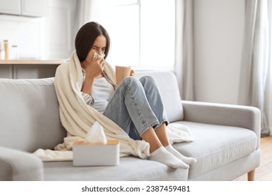 Cold And Flu Concept. Sick young woman blowing her running nose in paper tissue and drinking tea at home, ill female covered in blanket sitting on couch in living room, feeling unwell, copy space