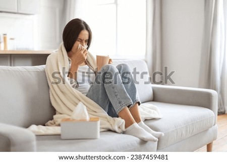 Cold And Flu Concept. Sick young woman blowing her running nose in paper tissue and drinking tea at home, ill female covered in blanket sitting on couch in living room, feeling unwell, copy space
