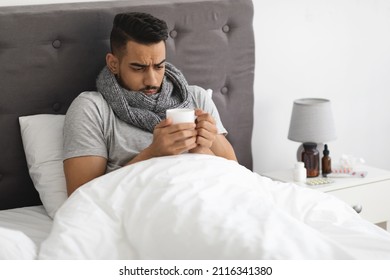 Cold And Flu. Portrait Of Sick Young Arab Man Drinking Tea In Bed, Ill Middle Eastern Guy Wearing Scarf Sitting With Cup Of Hot Drink In Hands, Having Fever, Feeling Unwell At Home, Closeup Shot