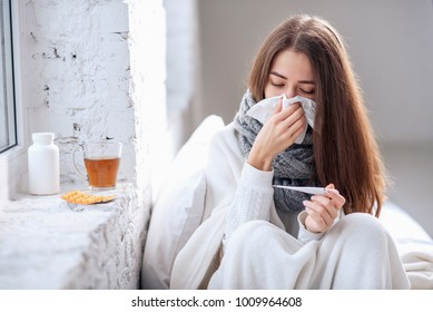 Cold and flu. Sick woman caught cold, feeling illness and sneezing in paper wipe. Closeup of beautiful unhealthy girl covered in blanket wiping nose and looking at thermometer. Healthcare concept. - Shutterstock ID 1009964608