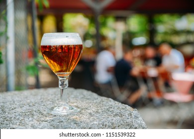 Cold Full Beer Glass On Granite Table With Busy Beer Garden Scene In The Defocused Background