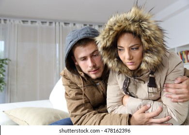 Cold home with an angry couple warmly clothed hugging sitting on a sofa in the living room