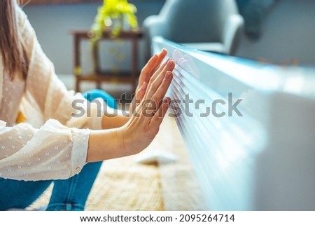 Cold home, freezing. Using heater at home in winter. Woman warming her hands sitting by device and wearing warm clothes. Heating season. Woman warms up hands over heater.