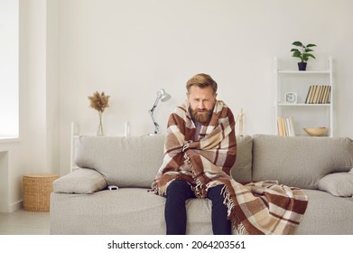 It's cold at home in wintertime. Man freezing in his house in winter because of broken thermostat. Sad bearded young guy wrapped in woolen plaid shivering while sitting on sofa in living room interior