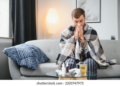 It's Cold At Home In Wintertime. Man Freezing In His House In Winter Because Of Broken Thermostat. Young Guy Wrapped In Woolen Plaid Shivering While Sitting On Sofa In Living Room Interior.