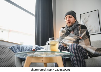 It's Cold At Home In Wintertime. Man Freezing In His House In Winter Because Of Broken Thermostat. Young Guy Wrapped In Woolen Plaid Shivering While Sitting On Sofa In Living Room Interior.