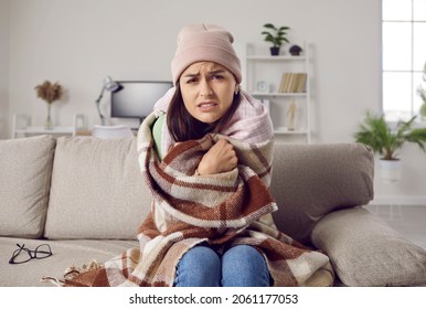 It's so cold in house in winter. Woman freezes in wintertime due to problems with central heating. Young girl wearing warm hat and wrapped in wool blanket sitting on sofa at home and looking at camera