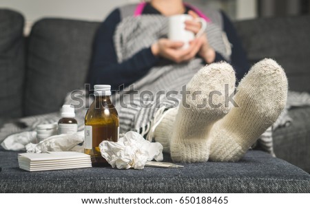 Cold medicine and sick woman drinking hot beverage to get well from flu, fever and virus. Dirty paper towels and tissues on table. Ill person wearing warm woolen stocking socks in winter. 