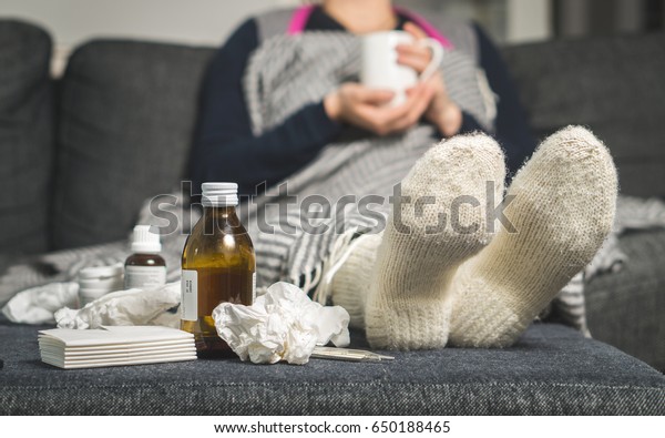 Cold medicine and sick woman drinking hot beverage to\
get well from flu, fever and virus. Dirty paper towels and tissues\
on table. Ill person wearing warm woolen stocking socks in winter.\
