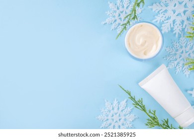 Cold protecting winter cream concept. Simple tubes and jar cosmetics mockup background with artificial snowflakes and fir tree branches, top view on light blue background. Winter seasonal skin care