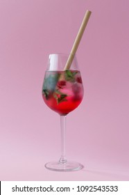 Cold refreshing drink with diffusing grenadine syrup, fresh mint leaves, raspberries and ice in a wineglass standing on a pastel pink background. It has a ecological bamboo straw in it.