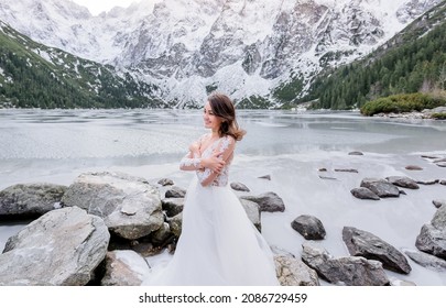 Cold Smiled Girl Dressed In The Wedding Dress Is Standing Near The Frozen Highland Lake In Winter