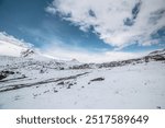 Cold snow-white landscape with snow-covered pointy peak under clouds in blue sky. Snowy rocky mountainside and mountain peaked top. Last year