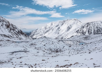 Cold snow-white landscape of snowy valley with alpine lakes against snow-covered rocky mountain range under clouds in blue sky. Glacial lakes, pure white snow and stones in high mountains in sunny day - Powered by Shutterstock