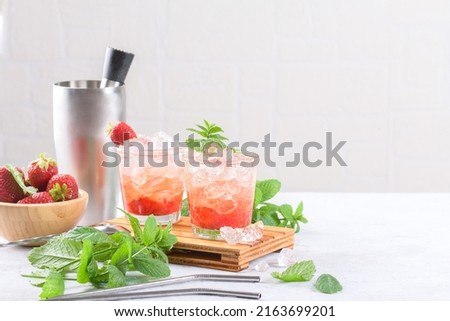 Cold summer strawberry cocktail mojito, margarita, daiquiri. Two glasses with fresh strawberry soda drink, ice cubes, mint and bartender tools on light background. Iced strawberry lemonade, copy space